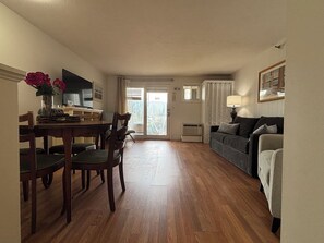 Ein Wohnzimmer mit einer Couch, einem Esstisch mit Stühlen, einem Fernseher und einem Fenster mit Vorhängen.