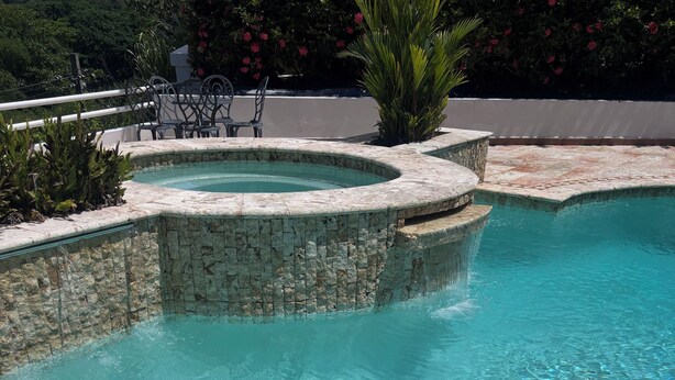 Une piscine aux eaux cristallines, bordée de pierres, entourée d’une terrasse aménagée avec des chaises et un petit jacuzzi.