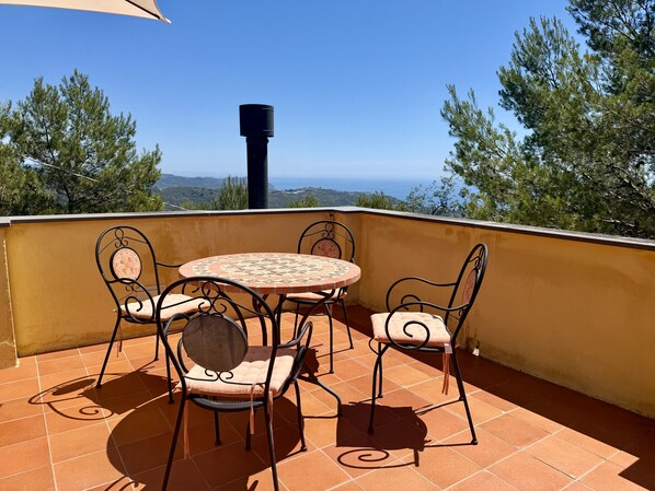 A terrace with a round table and four chairs, a chimney, and a view of the sea and trees.