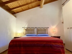 A bedroom with a wooden ceiling, a bed with a red and blue striped bedspread, a nightstand with a lamp, and a wardrobe.