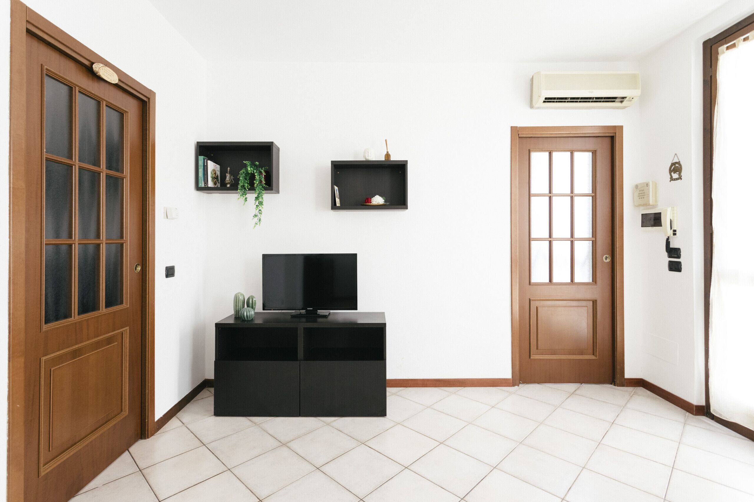 A room with a wooden door, a black entertainment unit with a TV, a small shelf with decorative items, and a white tiled floor.
