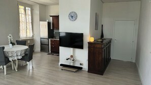 En matplats med ett bord och stolar, ett kök med vitvaror, en platt-TV som är monterad på väggen och en klocka.