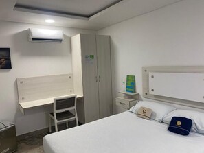 Et hotelværelse med seng, skrivebord, stol, aircondition, garderobeskab og et natbord med lampe.