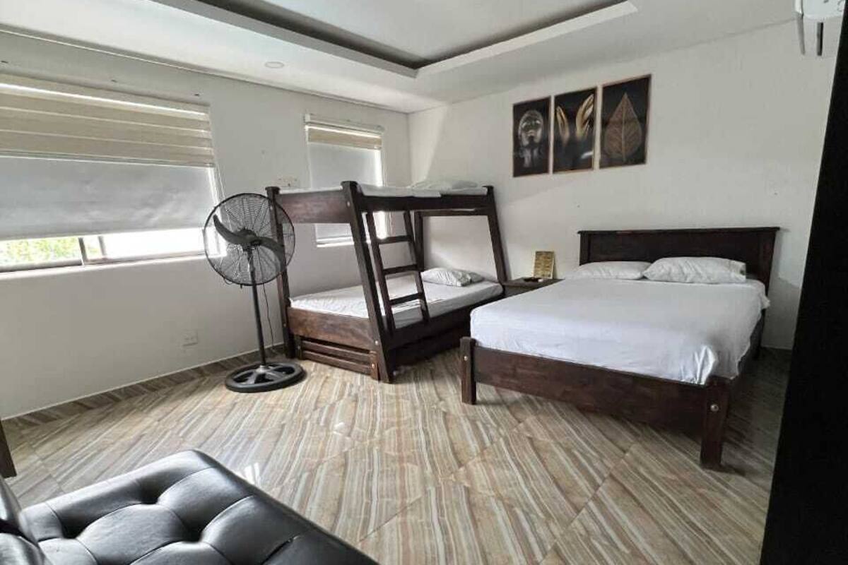 Ein Schlafzimmer mit einem Etagenbett, einem Einzelbett, einem schwarzen Ledersofa, einem Standventilator und einem Fenster mit Jalousien.