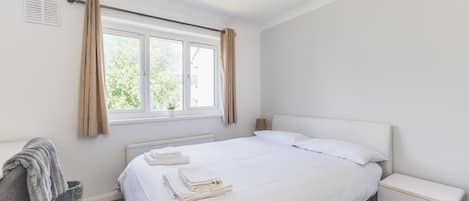 A hotel room with a bed, a bedside table, a radiator, a window with curtains, and a towel rack.