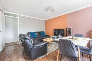 A living room with a sofa, a TV, a dining table with chairs, and a small side table. - Modern 2BDR Flat w Parking Near Heathrow and Tube (London)