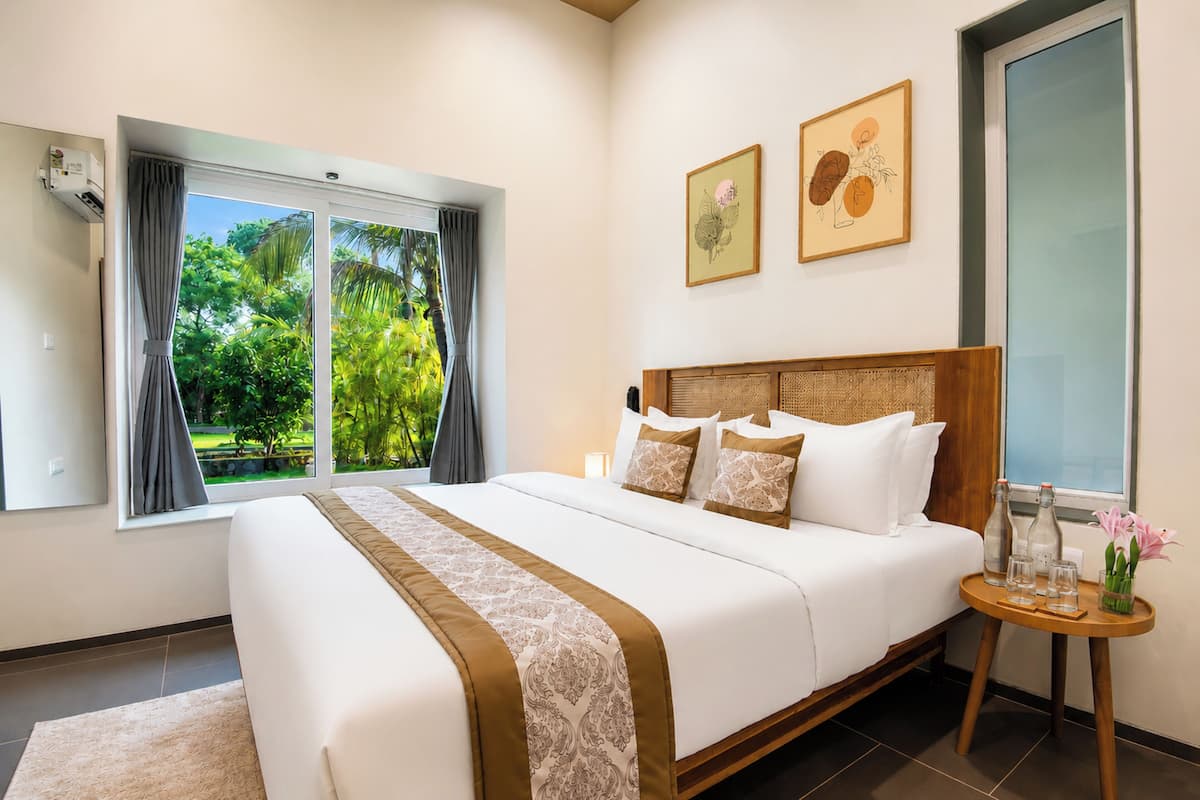 A bedroom with a large bed, a bedside table with glasses and a vase, and a window with curtains.