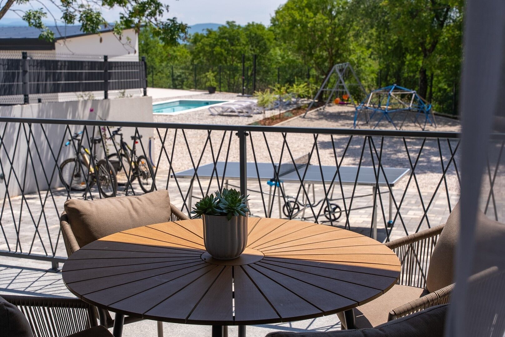 A round table with a potted plant, outdoor seating, a pool, and bicycles in the background.