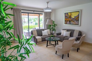Ein Wohnzimmer mit zwei Sofas, einem Glastisch und einer Lampe. An der Wand hängt ein Gemälde und im Vordergrund steht eine Pflanze.