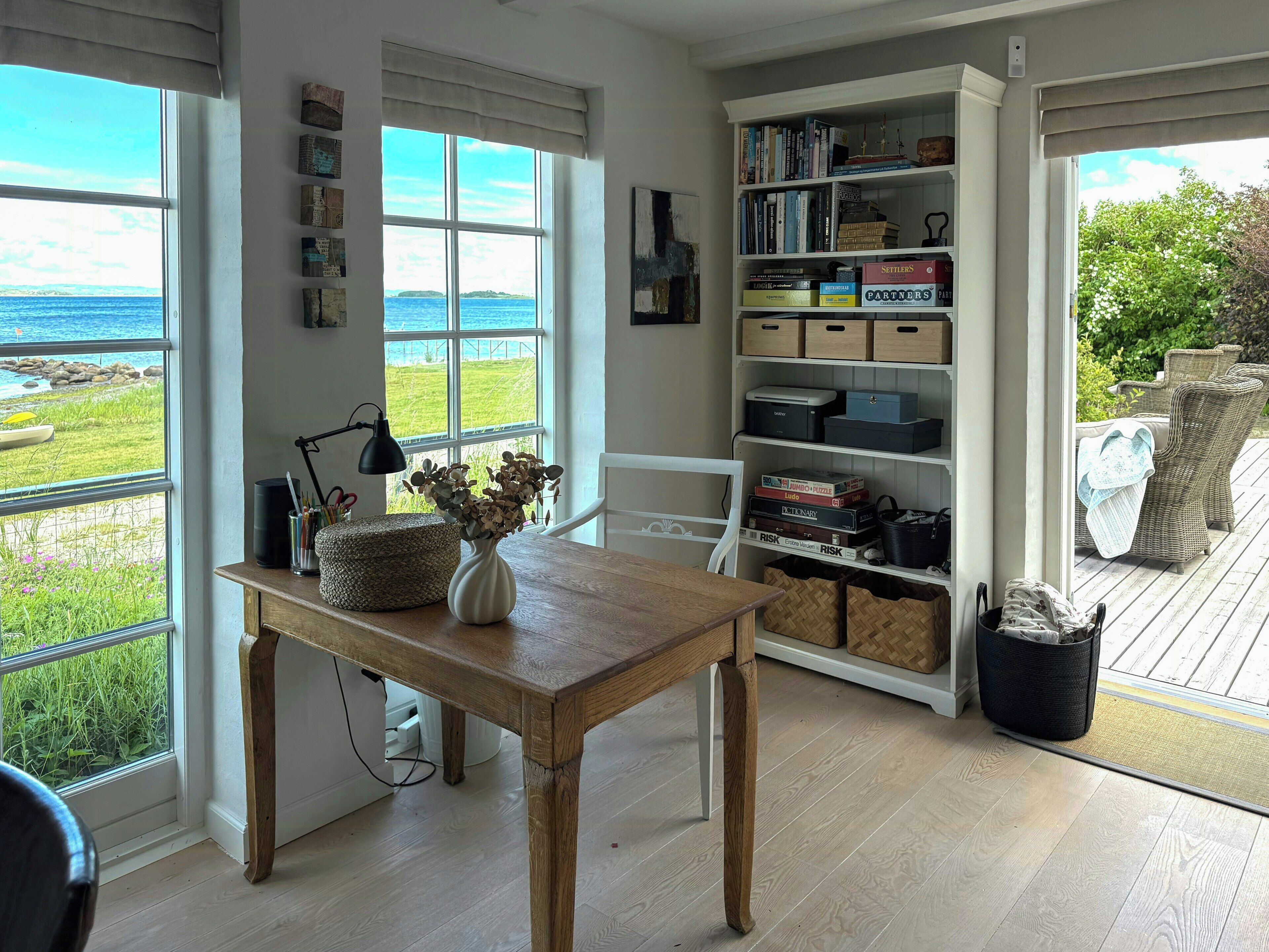 Una habitación con vista al mar, una mesa de madera con un florero, una estantería y muebles de mimbre.
