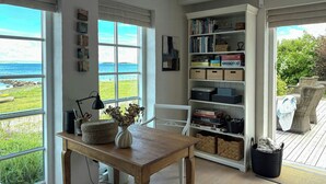 A room with a view of the sea, a wooden table with a vase, a bookshelf, and wicker furniture.