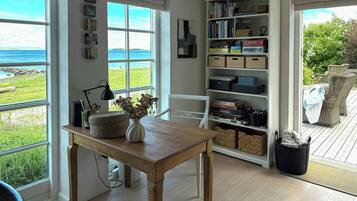 Una habitación con vista al mar, una mesa de madera con un florero, una estantería y muebles de mimbre.