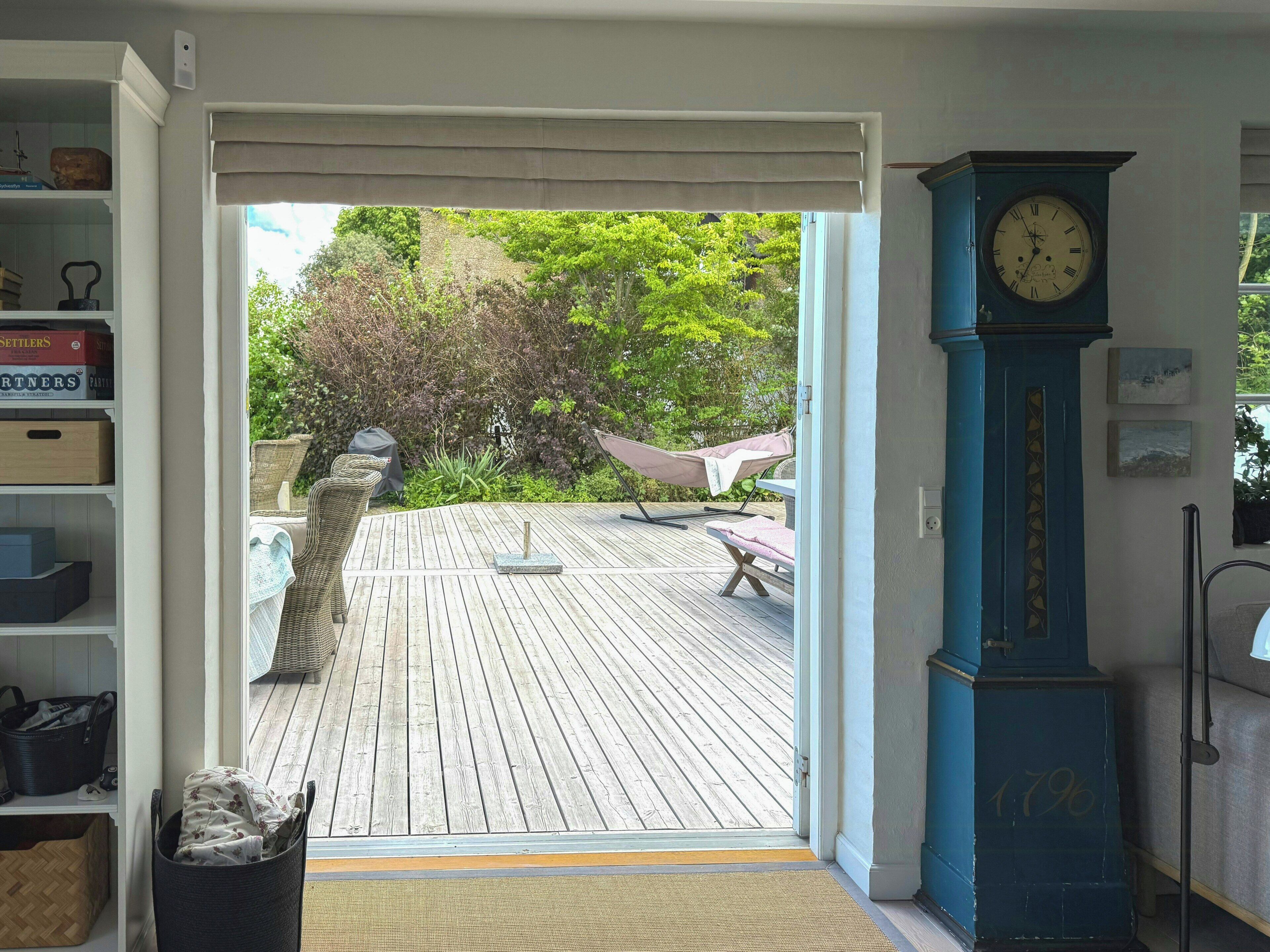 Ein offenes Durchgang zu einer Holzterrasse mit Gartenmöbeln, einer Uhr und einem Bücherregal mit Büchern.