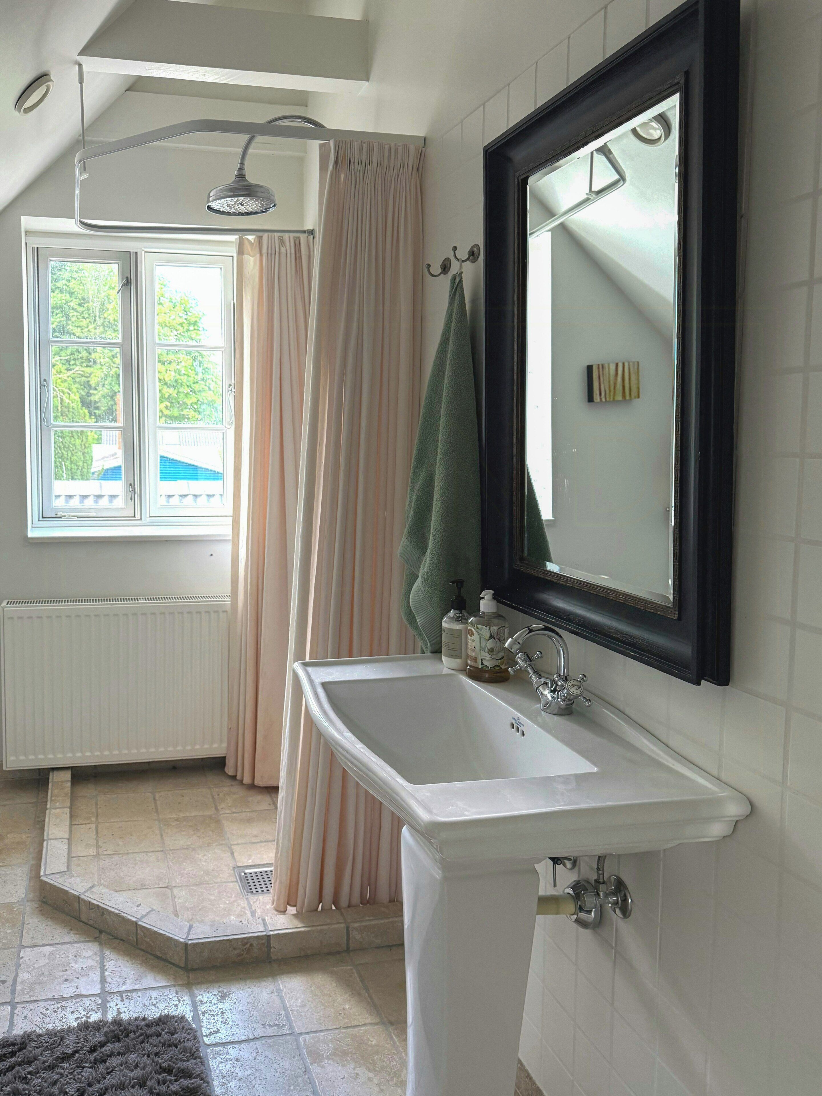 Un bany amb un lavabo blanc, un mirall, una dutxa i una finestra amb cortines.
