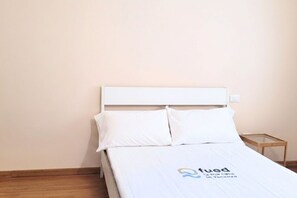 Ein Einzelbett mit weißer Bettwäsche und Kopfteil, ein kleiner Nachttisch daneben und eine schlichte Wand.