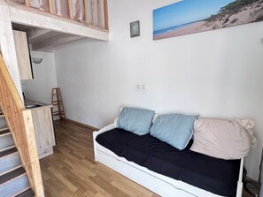 Een kamer met een houten trap, een witte bank met gekleurde kussens, een klein ladders en een foto met een strandthema aan de muur.