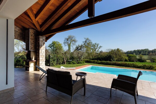 A covered patio with a stone fireplace, two wicker chairs, and a view of a swimming pool and surrounding greenery.
