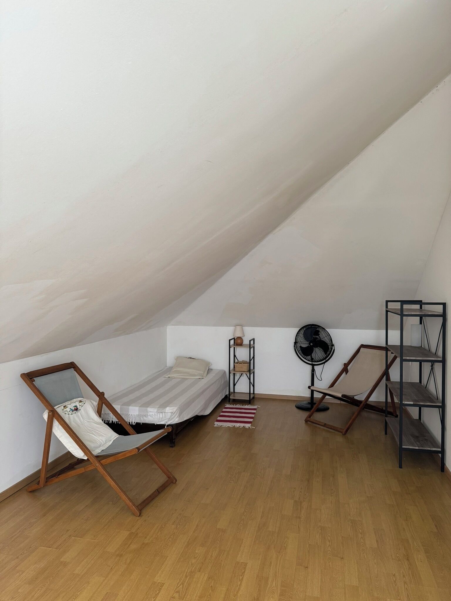 Une petite pièce avec un toit en pente, comprenant un lit simple, une chaise en bois, un ventilateur sur pied et une étagère en métal.