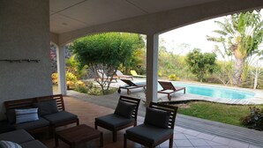 Eine überdachte Terrasse mit Gartenmöbeln, einem Swimmingpool und Blick auf den umliegenden Garten.