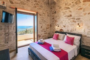 Ein Schlafzimmer mit Steinwand, einem Bett mit weißer Bettwäsche und pinken Kissen, einer roten Decke und Blick auf das Meer durch eine Glastür.