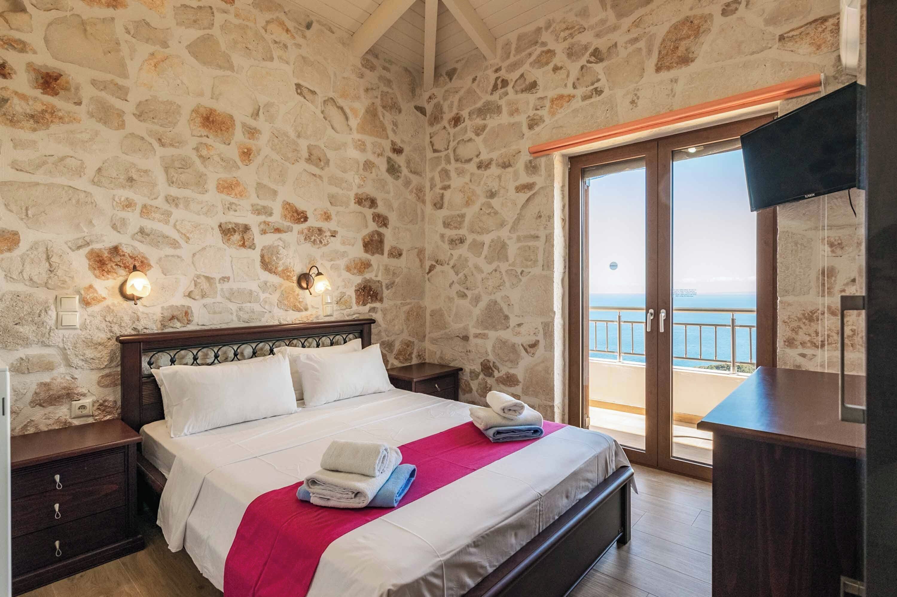 Een slaapkamer met een stenen muur, een bed met witte lakens, een rode deken en uitzicht op zee door een glazen deur.