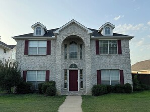 A two-story house with a brick facade, a central entrance with a red door, and a small front yard. - Spacious 4 Bedroom home with private backyard (Irving)