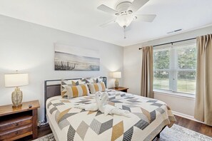 Un dormitorio con cama, mesitas de noche, ventilador de techo, un cuadro y una ventana con cortinas.
