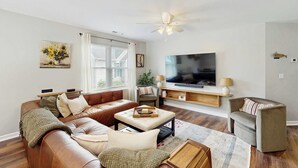 Ein Wohnzimmer mit braunem Ledersofa, einem beige-farbenen Hocker, einem grauen Sessel, einem Flachbildfernseher und einem Deckenventilator.