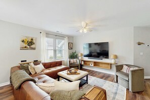 A living room with a brown leather sofa, a beige ottoman, a grey armchair, a flat-screen TV, and a ceiling fan. - Rehoboth Beach - 1 Melody Ct (Rehoboth Beach)
