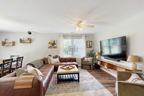 Ein Wohnzimmer mit Sofa, Hocker, Fernseher, Deckenventilator und einem Essbereich mit Stühlen und Tisch.