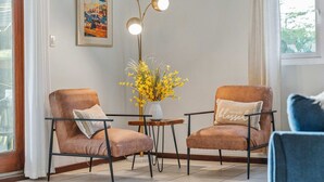 Um quarto com duas poltronas marrons, uma mesa pequena com um vaso de flores amarelas, um quadro na parede e um abajur com três lâmpadas.