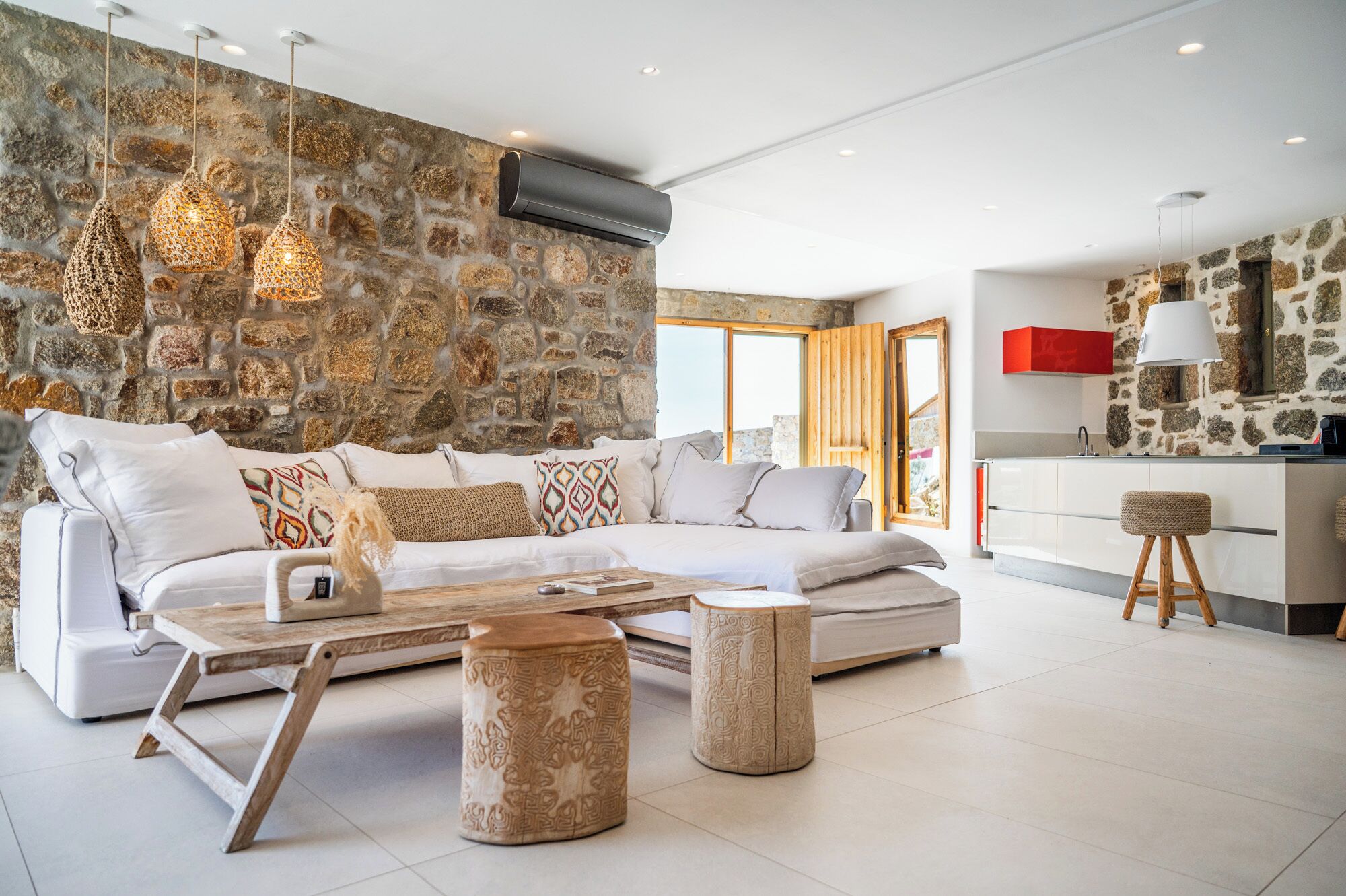 A modern living room with a stone wall, a white sofa, patterned cushions, a coffee table, and a kitchen area with a red cabinet and a hanging light fixture.