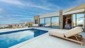A modern pool area with a clear view of the sea, a lounge chair, and a stone wall. - Citadel Natural Stone Villa (Mykonos)