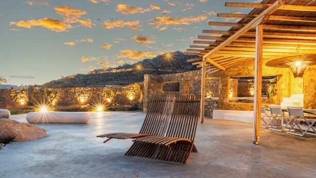 A sunlit outdoor seating area with a stone wall backdrop, a wooden pergola, and a stone structure with a fire feature. - Citadel Natural Stone Villa (Mykonos)