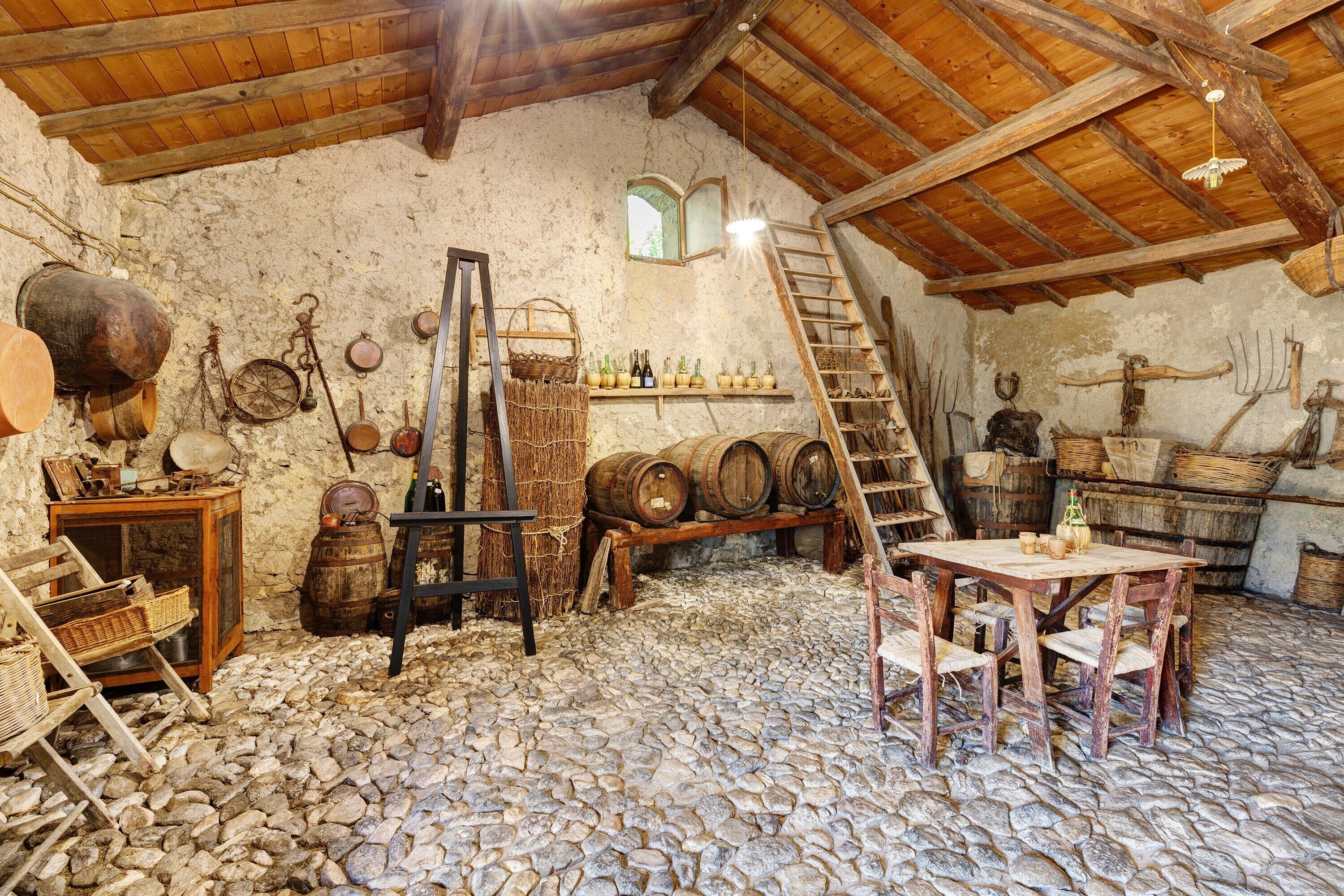 A rustic room with stone flooring, wooden furniture, and various tools and barrels.
