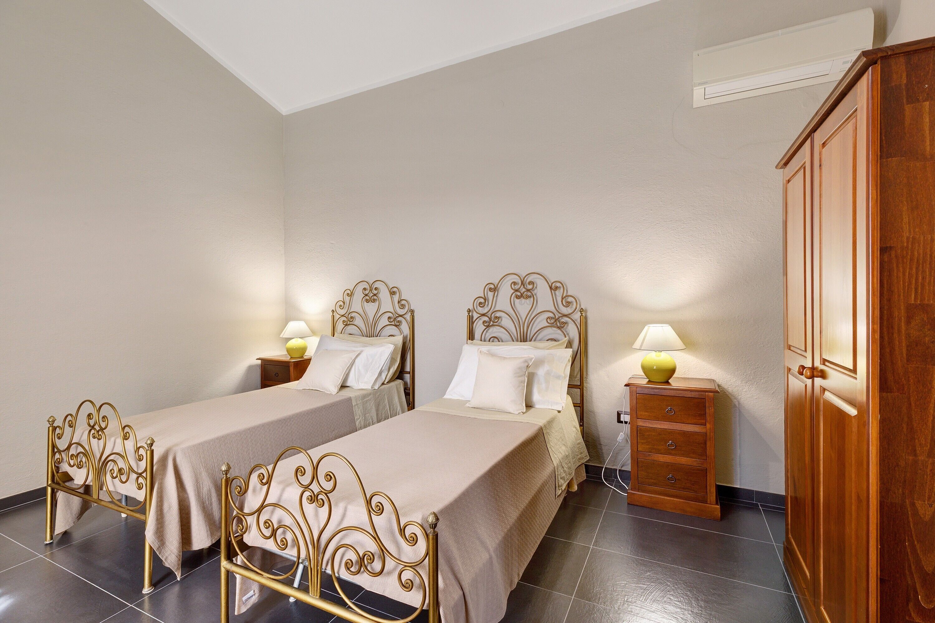 Quarto com duas camas de solteiro, guarda-roupa de madeira, mesa de cabeceira com abajur e piso de azulejos.
