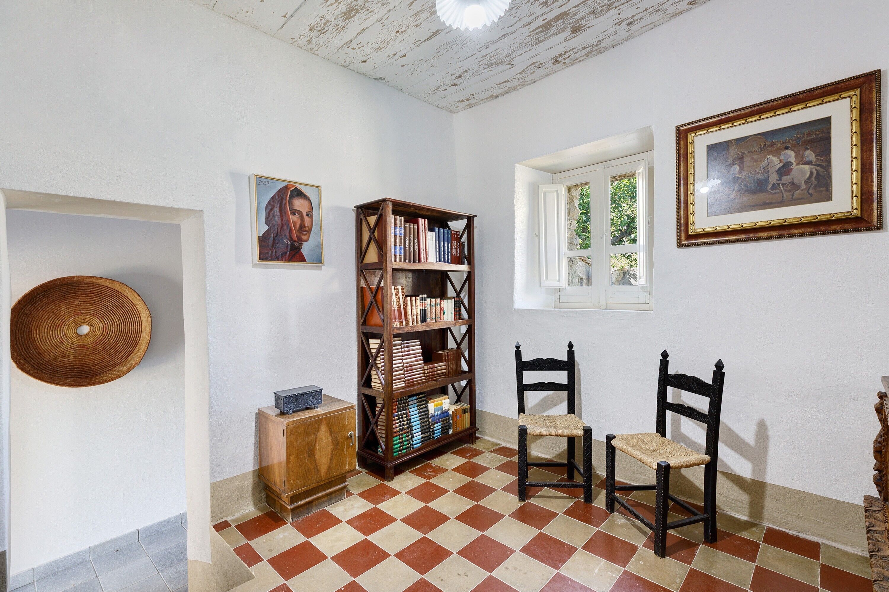 Una stanza con pavimento a scacchi, una libreria in legno, due sedie, un piccolo mobile e un quadro appeso al muro.