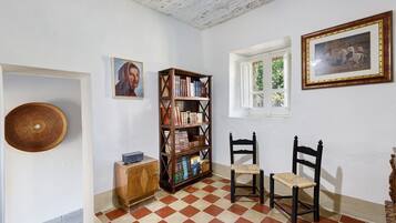 Una stanza con pavimento a scacchi, una libreria in legno, due sedie, un piccolo mobile e un quadro appeso al muro.