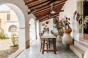 A covered outdoor dining area with a rustic wooden table and chairs, potted plants, and a view of the street.