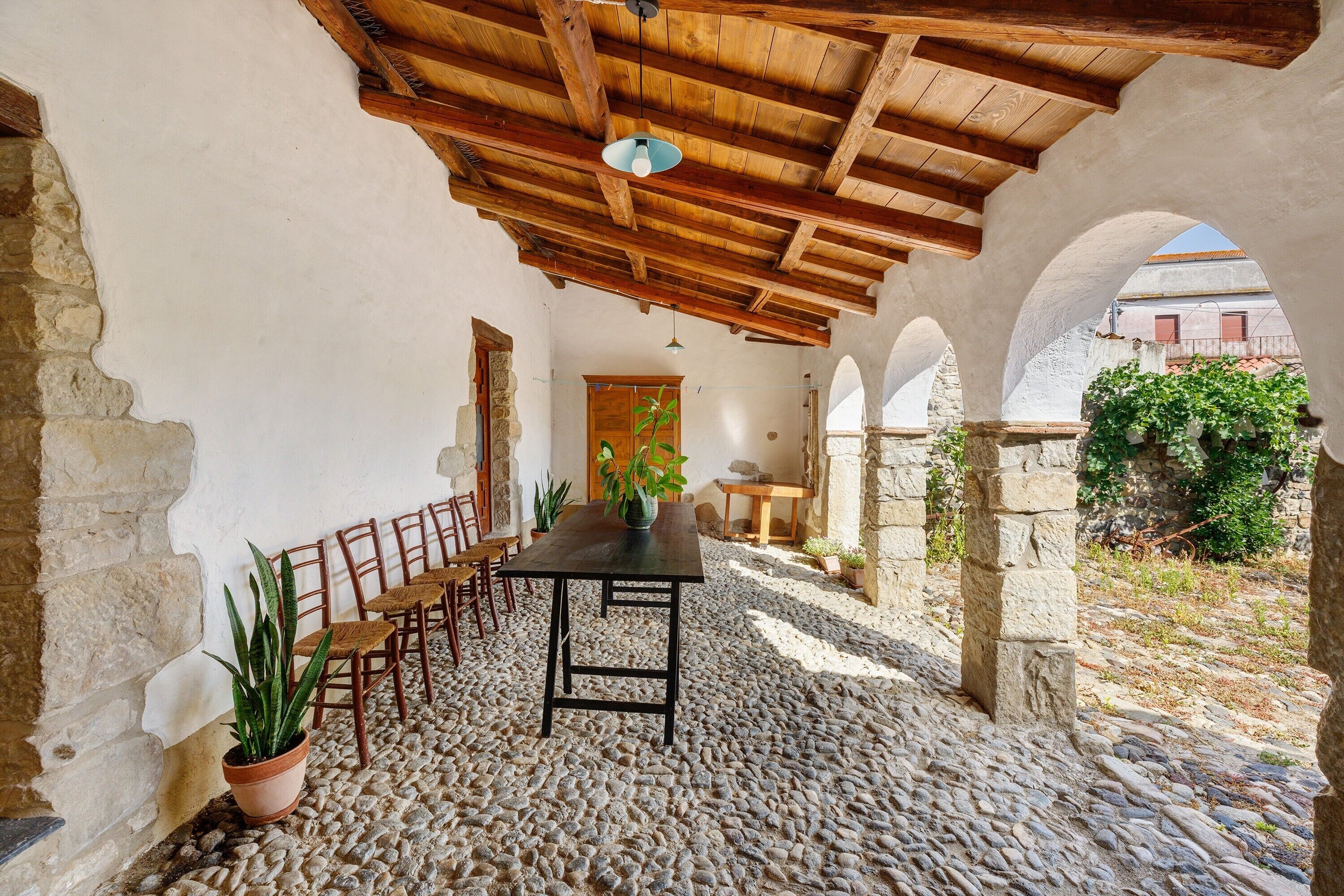 A rustic stone patio with a wooden table and chairs, surrounded by arches and a wooden ceiling.