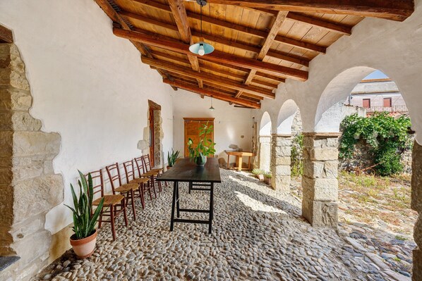 A rustic stone patio with a wooden table and chairs, surrounded by arches and a wooden ceiling.