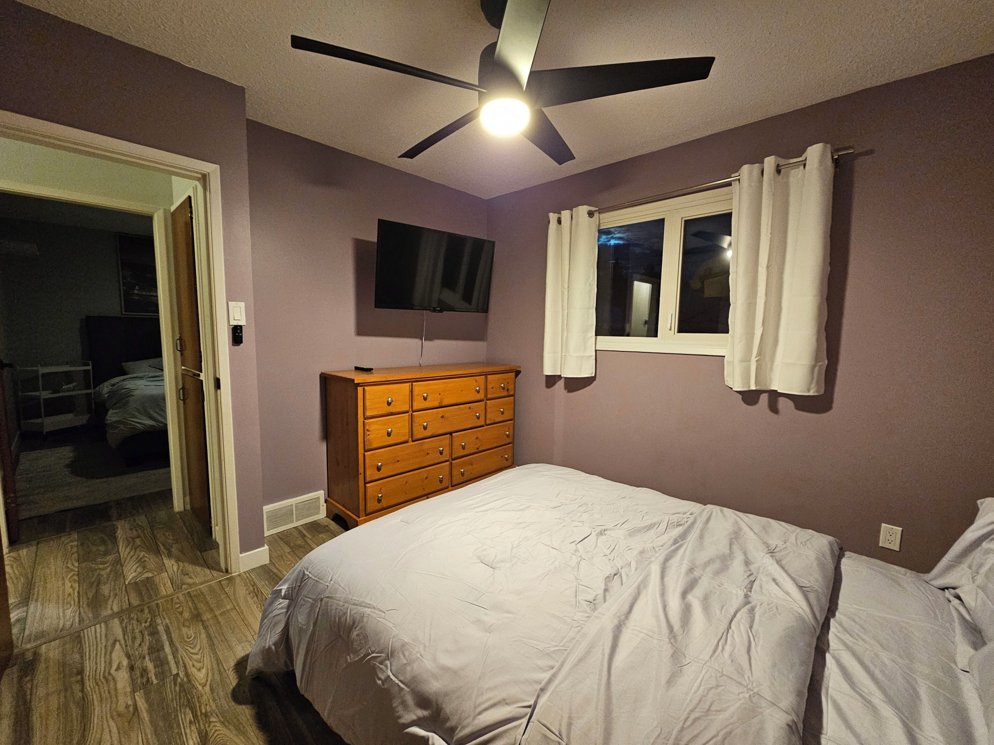 A bedroom with a bed, a dresser, a ceiling fan, a TV, and a window with curtains.