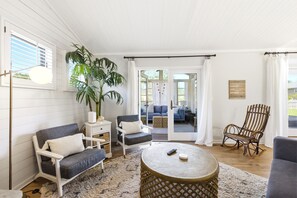 Une pièce avec un mur blanc, une grande plante en pot, un canapé gris, une table ronde et un fauteuil à bascule en bois.