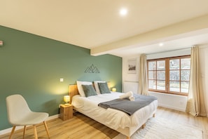 Ein Schlafzimmer mit einem Bett, einem Sessel, einem Nachttisch, einer Wandlampe und einem Fenster mit Vorhängen.