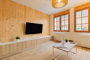 Ein Zimmer mit Holzwandverkleidung, einem an der Wand befestigten Flachbildfernseher, einer weißen Bank, einem kleinen Tisch mit einer Zeitschrift und großen Fenstern mit Blick ins Freie.