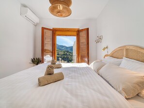 A bedroom with a large bed, a rattan headboard, a ceiling light, and a view of mountains through an open door. - Casa Lavender Frigiliana - A peaceful retreat bathed in light (Frigiliana)