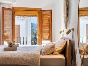 A bedroom with a bed, a sofa, a balcony with wooden doors, and a view of a mountainous landscape. - Casa Lavender Frigiliana - A peaceful retreat bathed in light (Frigiliana)