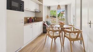 Une cuisine moderne avec des meubles blancs et un micro-ondes noir, un coin repas avec des chaises en bois et une table, et une vue sur un jardin à travers une porte vitrée.
