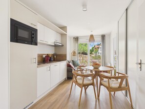 A modern kitchen with white cabinets and a black microwave, a dining area with wooden chairs and a table, and a view of a garden through a glass door. - The Swallow - Apt for 4 - Shared Pool (Biscarrosse)
