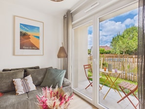 A living room with a sofa, pillows, a framed picture, and a view of the outdoors through a sliding glass door. - The Swallow - Apt for 4 - Shared Pool (Biscarrosse)